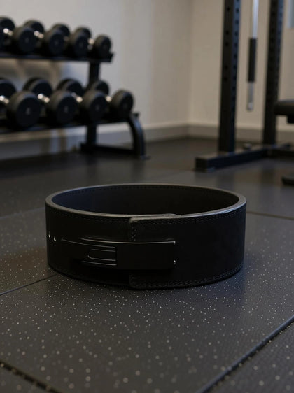 Foto real del Cinturón Forge apoyado en el piso de un gimnasio, con mancuernas y rack de fondo